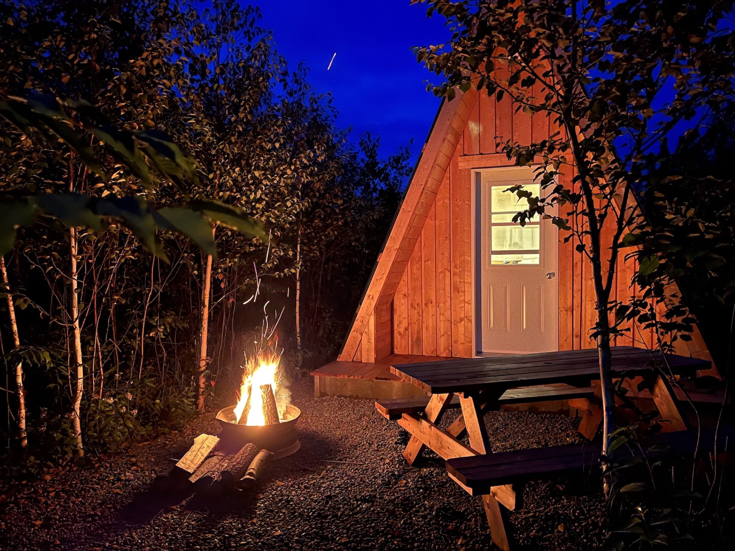 Mini-chalet prêt-à-camper en forêt boréale au crépuscule, avec feu de camp et table à pique-nique